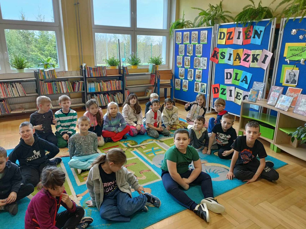 Uczniowie siedzą na dywanie, na tle gazetki o książkach dla dzieci.