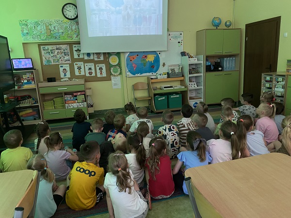 Dzieci siedzą na dywanie odwrócone do ekranu, oglądają film edukacyjny.