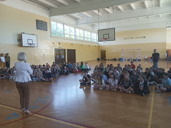 Pani dyrektor Małgorzata Fryt mówi do dzieci siedzących po turecku w dużej sali, życzy uczniom bezpiecznych wakacji