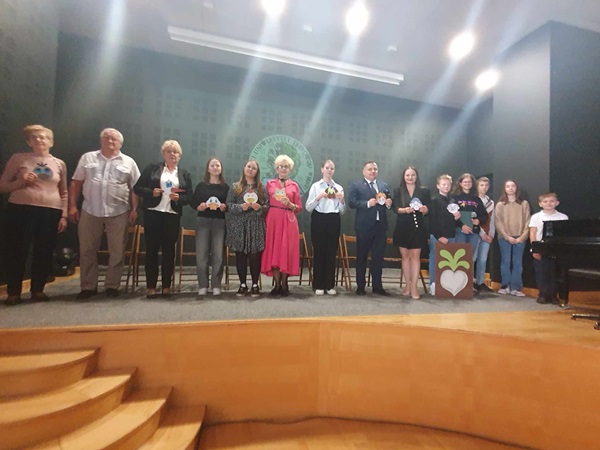 Na scenie stoją uczniowie i dorośli. Wszyscy kłaniają się po występie. Na ścianie wyświetlony jest logo Uniwersytetu Trzeciego Wieku