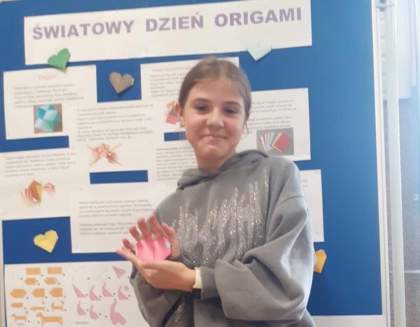 uczennica trzyma w ręku serduszko orgiami. Stoi na tle gazetki przedstawiającej historię sztuki składania papieru.