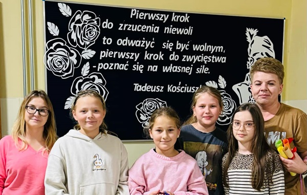 cztery uśmiechnięte dziewczynki, jedna trzyma koszyk wypełniony listami, druga medale dla nauczycieli, z dziećmi stoją dwie panie