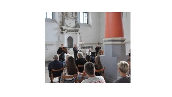 grupa ludzi siedzących na krzesłach, ujętych plecami, wysłuchuje koncertu czterech muzyków, ubranych na czarno we wnętrzu synagogi