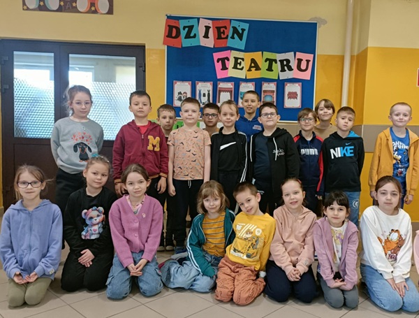 Dzieci z klasy 1C, stoją pod napisem: Dzień Teatru przy wejściu do biblioteki.