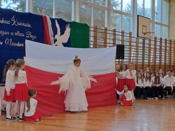 Dziewczynki tworzą flagę Polski z szarf, na jej tle stoi dziewczynka w przebraniu orła