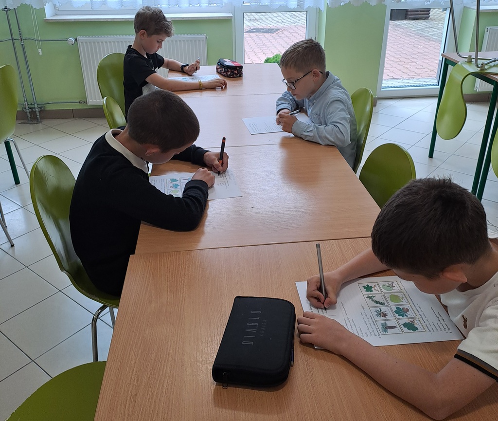Przedstawia dzieci siedzące przy stole i rozwiązujące test o lesie.