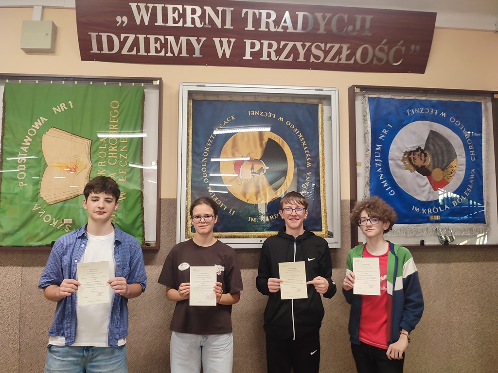 czworo uczniów stoi w rzędzie na tle ściany szkolnej. Każda z osób trzyma w rękach dyplom.