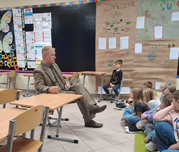 Leśniczy siedzący przy stole i rozmawiający z dziećmi.