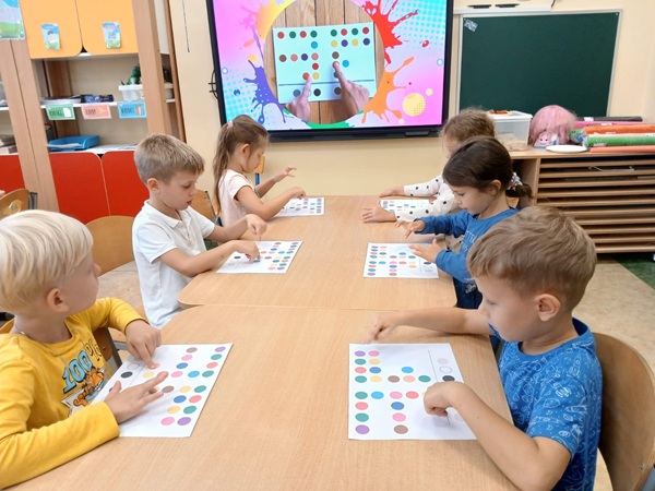 Dzieci siedzą przy stoliku, przed mini karta pracy, spoglądają na ekran multimedialny.