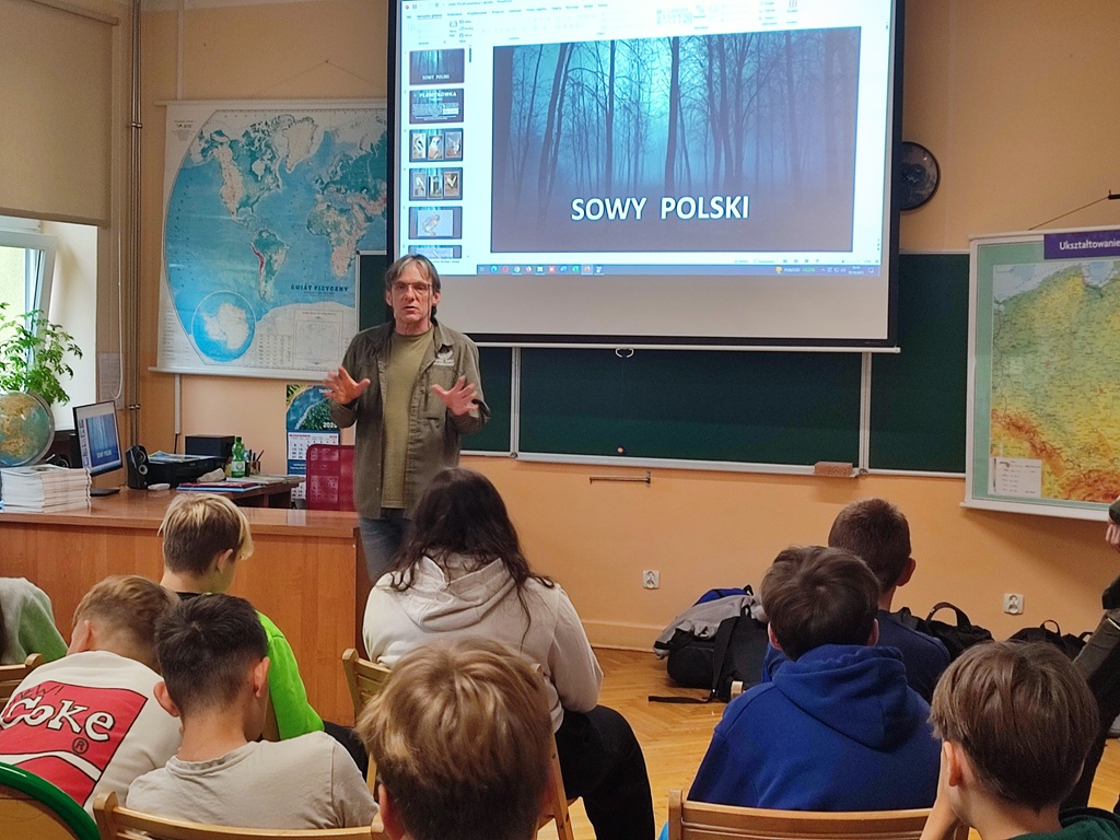 Sala lekcyjna z uczniami siedzącymi na krzesełkach na warsztatach. Na środku sali stoi mężczyzna.