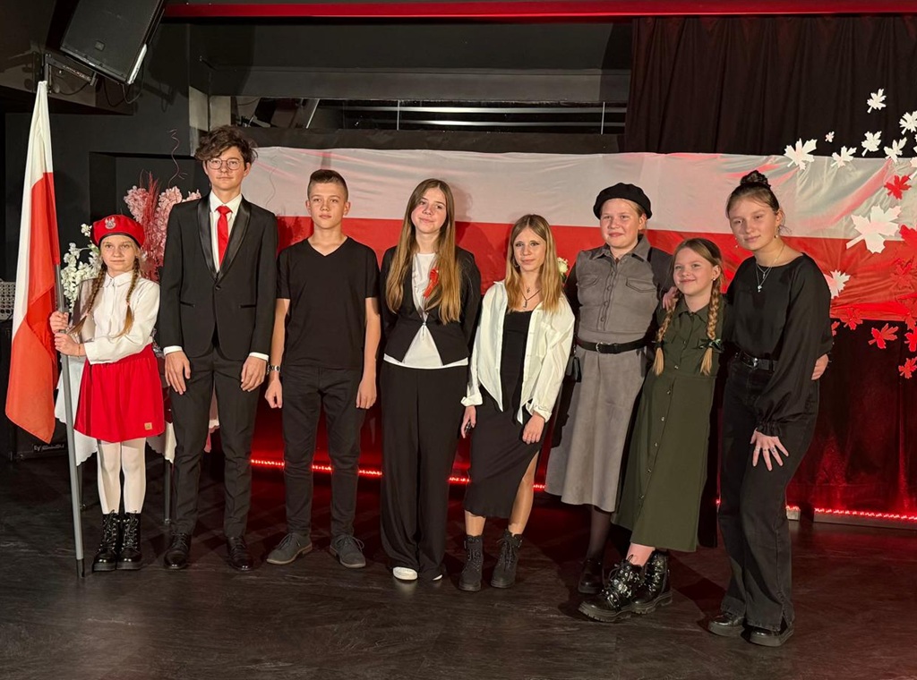 8 dzieci stojących w rzędzie. W tle dekoracja – flaga Polski i unoszące się z niej biało - czerwone liście z papieru