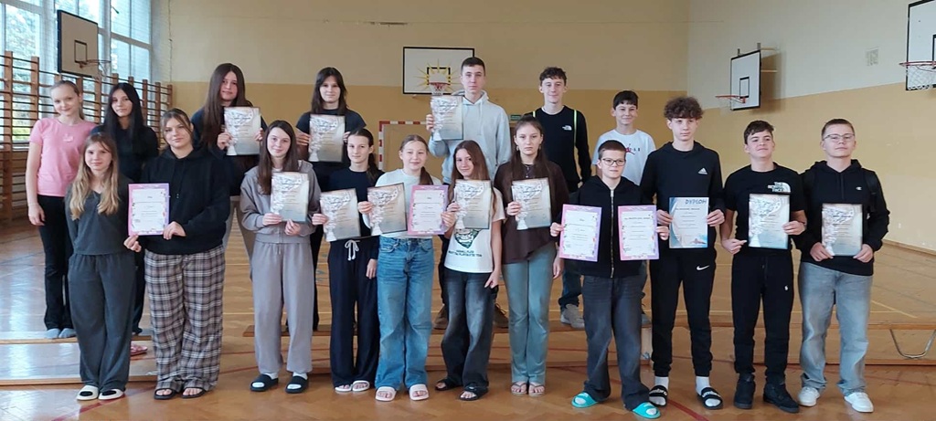 grupa uczniów stojąca w szkolnej sali gimnastycznej, dumnie trzymająca dyplomy.