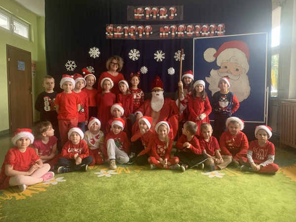 Grupa dzieci stoi na tle świątecznej dekoracji, pośrodku, na fotelu, siedzi Mikołaj.