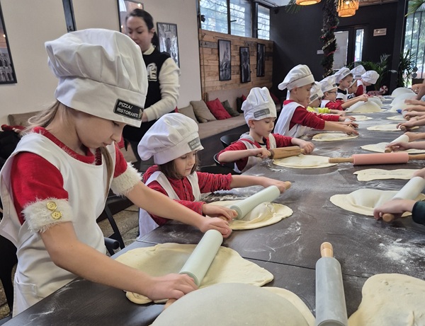 Dzieci w czapkach kucharskich z zaangażowaniem przygotowują ciasto na pizzę.