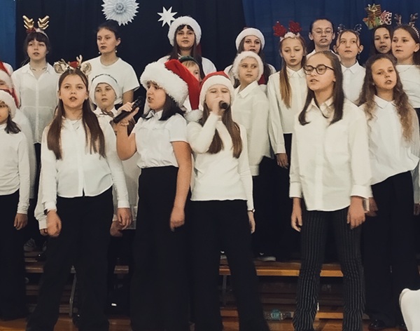 Dzieci śpiewające w szkolnym chórze, na niebieskim tle i świątecznej dekoracji.