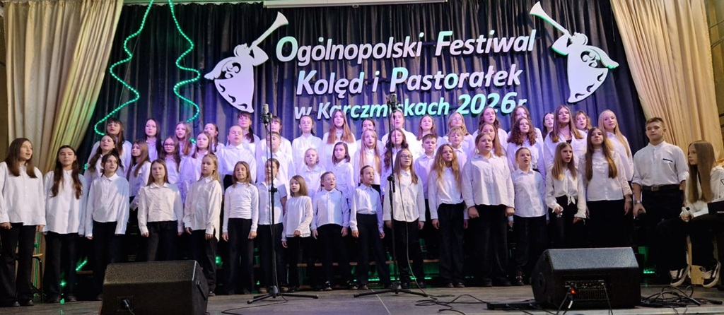 Chórzyści (ok. 50osób) stoją na scenie w białych koszulach i czarnych spodniach.