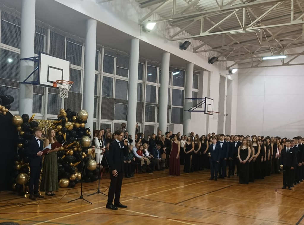 Chłopka trzyma mikrofon w oddali ósmoklasiści ustawieni w rzędach