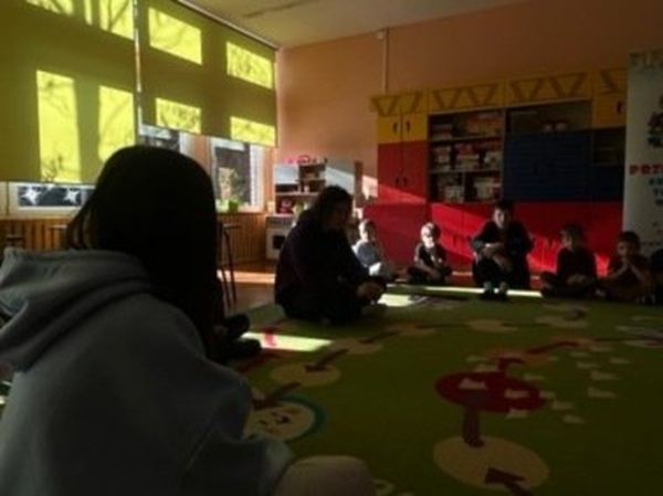 uczniowie i przedszkolaki siedzą na dywanie i słuchają czytania.