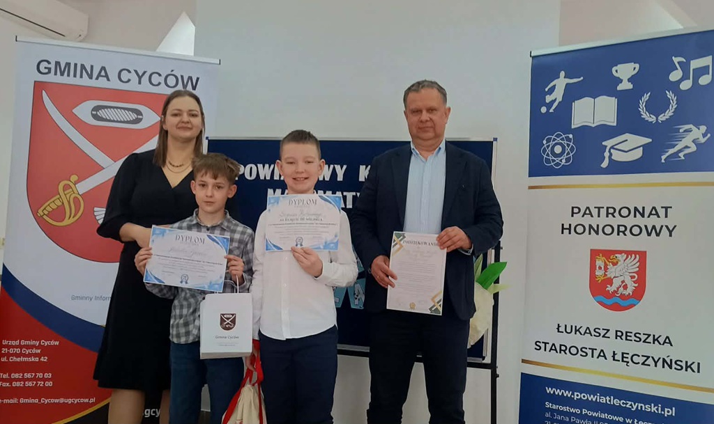 dwóch chłopców z dyplomami, obok osoby dorosłe, w tle tablice z logo konkursu