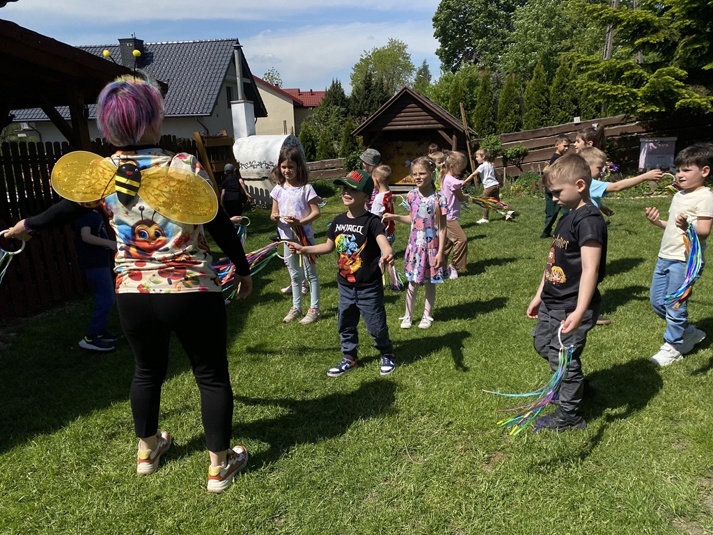 Dzieci z kolorowymi wstążkami biegają po trawniku obok kobiety w koszulce ze skrzydełkami pszczoły.