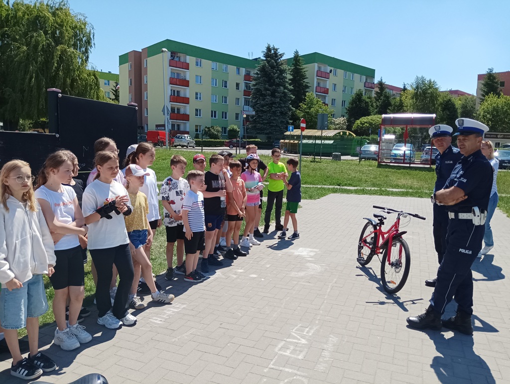 Uśmiechnięte dzieci stoją na chodniku, i słuchają wypowiedzi policjanta.