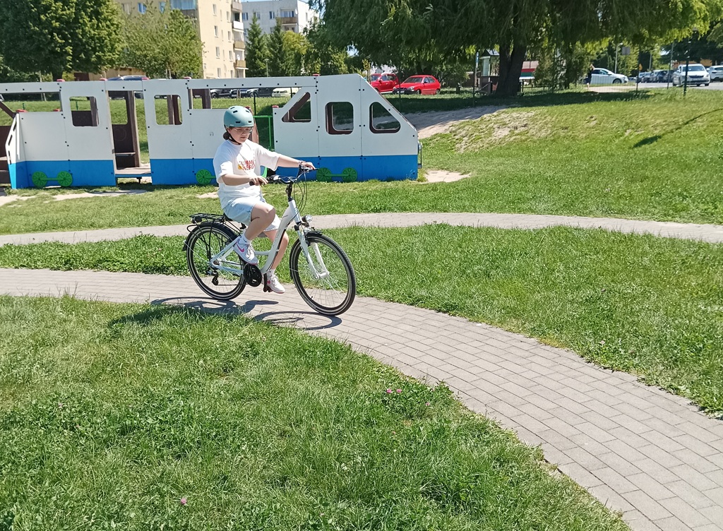 Dziewczynka w niebieskim kasku, jedzie na białym rowerze po chodniku.