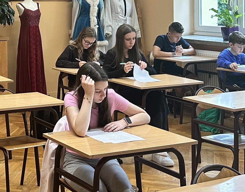 Uczniowie siedzą w ławkach w sali lekcyjnej i rozwiązują test na kartkach