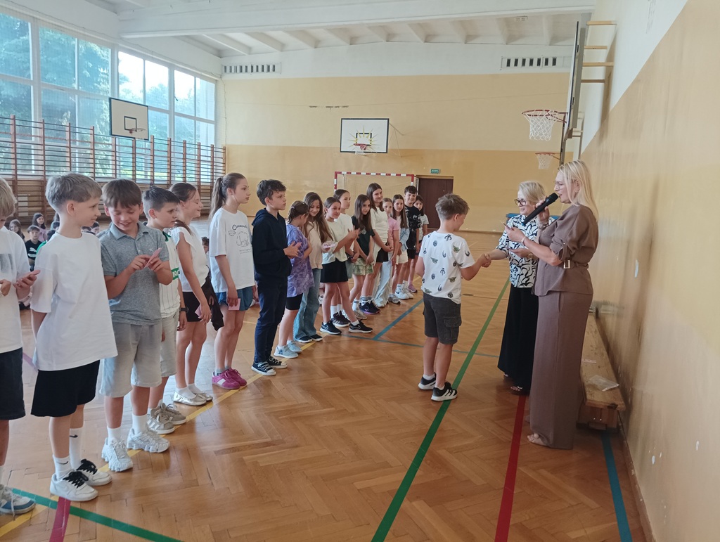 Pani dyrektor wręcza chłopcu kartę rowerową, zadowolone dzieci stoją na żółtej linii.