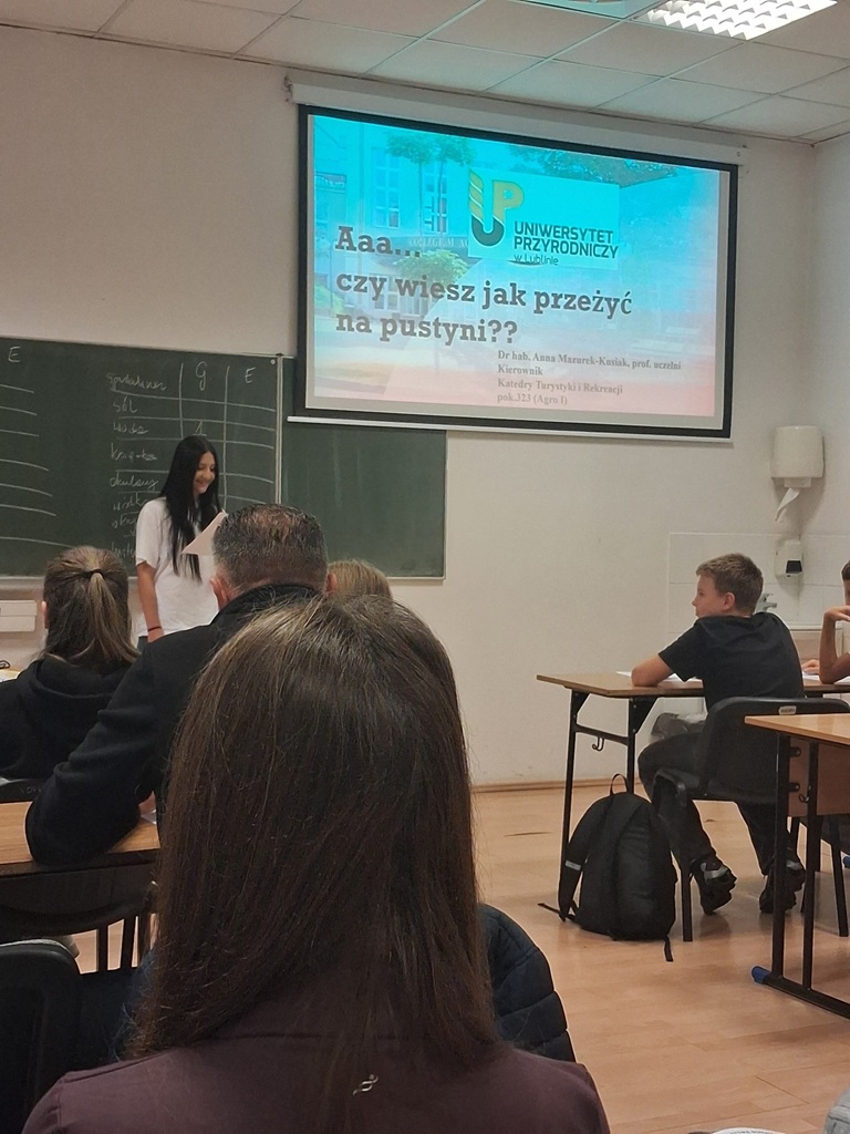 Uczennica w sali odczytuje z kartki informacje dla grupy uczniów siedzących w ławce.