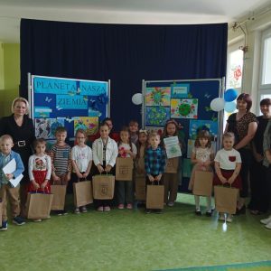 Dzieci stoją uśmiechnięte w ręku trzymają prezenty, w tle tablice z pracami konkursowymi.