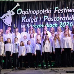 Chórzyści (ok. 50osób) stoją na scenie w białych koszulach i czarnych spodniach.