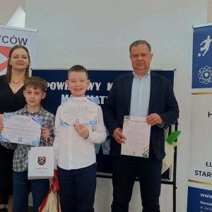 dwóch chłopców z dyplomami, obok osoby dorosłe, w tle tablice z logo konkursu