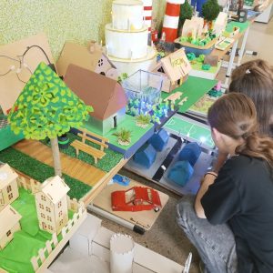widać trzy dziewczyny, które oglądają makietę z samochodami, osiedle niebieskich domków i boisko szkolne