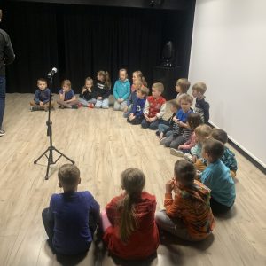 Dzieci siedzą na podłodze w kręgu, przed nimi stoi mikrofon.