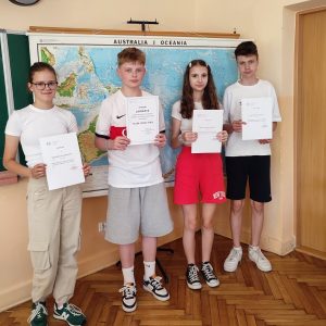 Czterech uczniów trzyma dyplomy, w tle z mapa ścienna Australia i Oceania.
