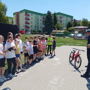 Uśmiechnięte dzieci stoją na chodniku, i słuchają wypowiedzi policjanta.