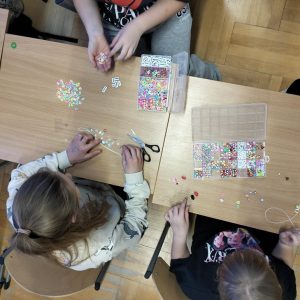 Dziewczynki siedzą przy stolikach robiąc zawieszki z koralików.