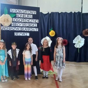Grupowe zdjęcie uczestników konkursu, w tle napis i papierowe, kolorowe kwiaty.