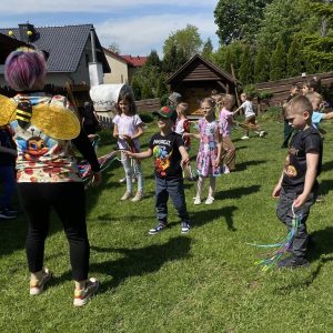 Dzieci z kolorowymi wstążkami biegają po trawniku obok kobiety w koszulce ze skrzydełkami pszczoły.