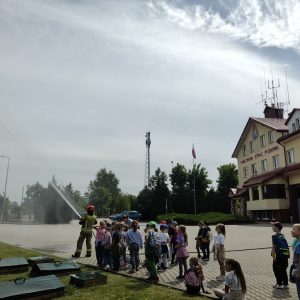 Grupa dzieci, słucha strażaków, w tle wozy strażackie