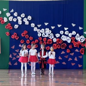 Trzy dziewczynki w białych bluzkach i czerwonych spódnicach i chłopiec w stroju krakowskim recytują wiersze.