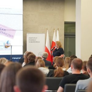 uczestnicy konferencji wysłuchują wystąpienia Wioletty Woś, prelegentka stoi z mikrofonem na tle ekranu