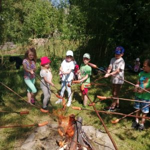 Dzieci uśmiechnięte stoją na trawie trzymają w ręku kij z kiełbaską nad ogniskiem.