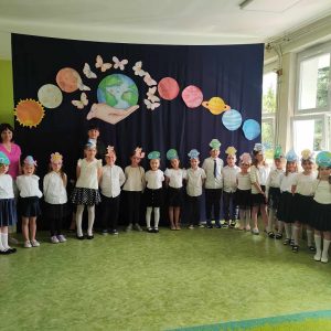 Grupa dzieci z nauczycielami stoi na korytarzu szkolnym, w tle dekoracja przedstawiająca planetę ziemię oraz inne planety układu słonecznego.