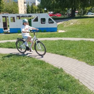 Dziewczynka w niebieskim kasku, jedzie na białym rowerze po chodniku.