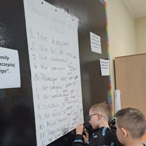Dwóch chłopców, którzy piszą na kartonie zawieszonym na ścianie.