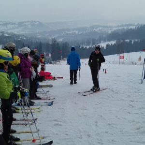 Dzieci ubrane w stroje narciarskie stoją przed instruktorem, w tle ośnieżone góry