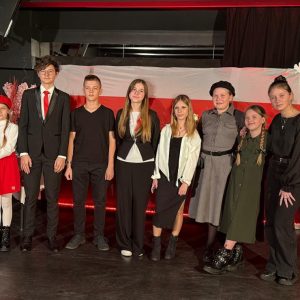 8 dzieci stojących w rzędzie. W tle dekoracja – flaga Polski i unoszące się z niej biało - czerwone liście z papieru