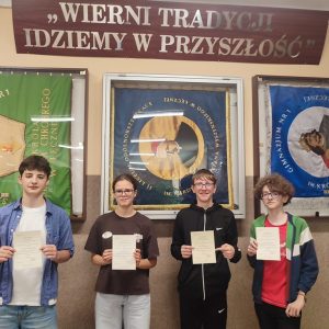czworo uczniów stoi w rzędzie na tle ściany szkolnej. Każda z osób trzyma w rękach dyplom.