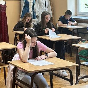 Uczniowie siedzą w ławkach w sali lekcyjnej i rozwiązują test na kartkach